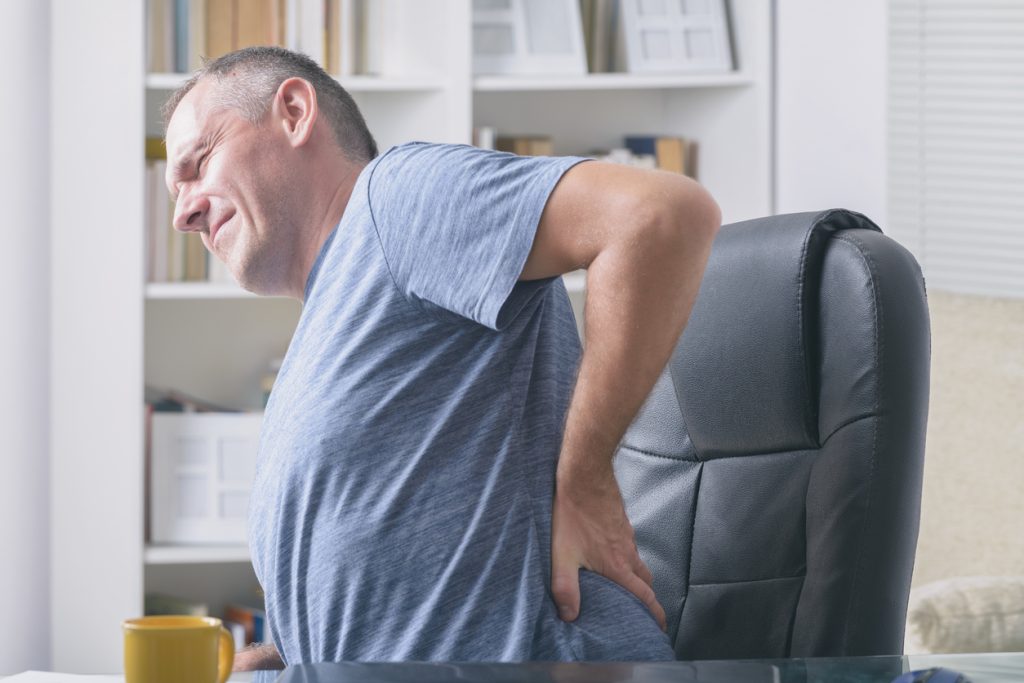 Man in home office suffering from sciatica and low back pain sitting at a desk with notebook, papers and other objects
